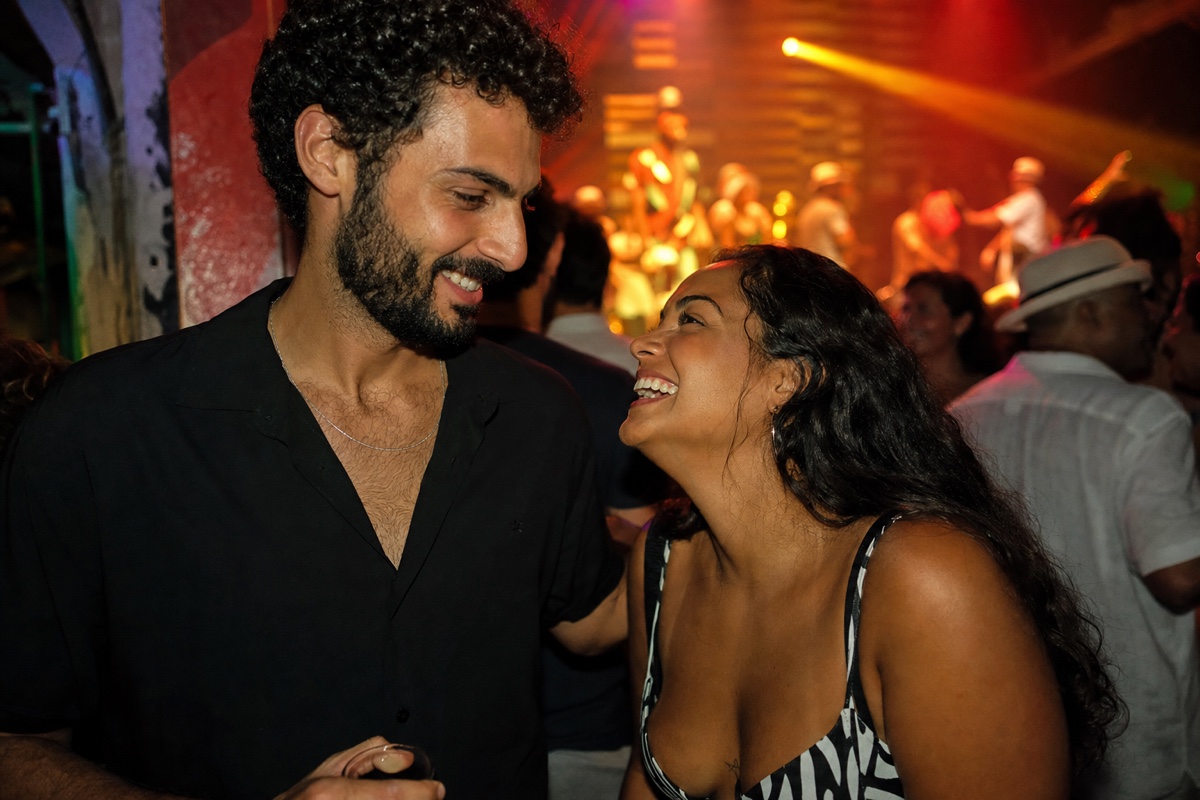 A Middle Eastern man and a Brazilian woman flirting at a lively Lapa nightclub in Rio, a samba crowd and warm stage lights behind them