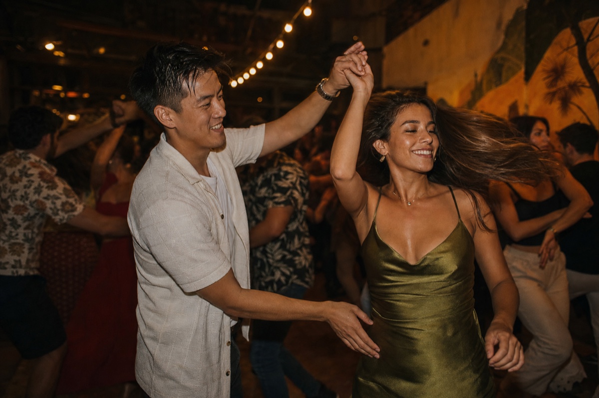 A couple dancing salsa in a warmly lit Cali club, an East Asian man and a Colombian woman mid-turn