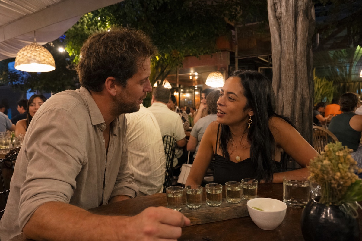A European man and a Mexican woman sharing a flight of mezcal at a warmly lit Roma Norte terrace, other tables full behind them